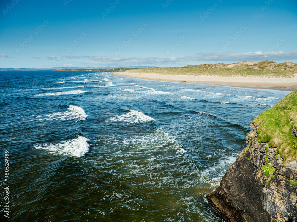 Fototapeta premium Green grass grows on cliff and amazing waves and sandy Bundoran beach in Ireland on warm sunny day. Popular tourist and surfers area. Blue cloudy sky. Irish landscape. Rich saturated color.
