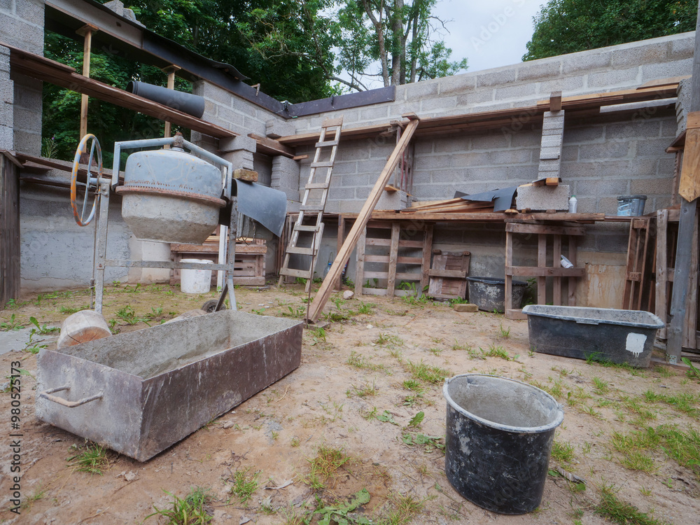 A construction site with a cement mixer inside. Scene is that of a work in progress, with the unfinished building and the tools scattered around and wooden walk way without safety. Small size home A construction site with a cement mixer inside. Scene is that of a work in progress, with the unfinished building and the tools scattered around and wooden walk way without safety. Small size home