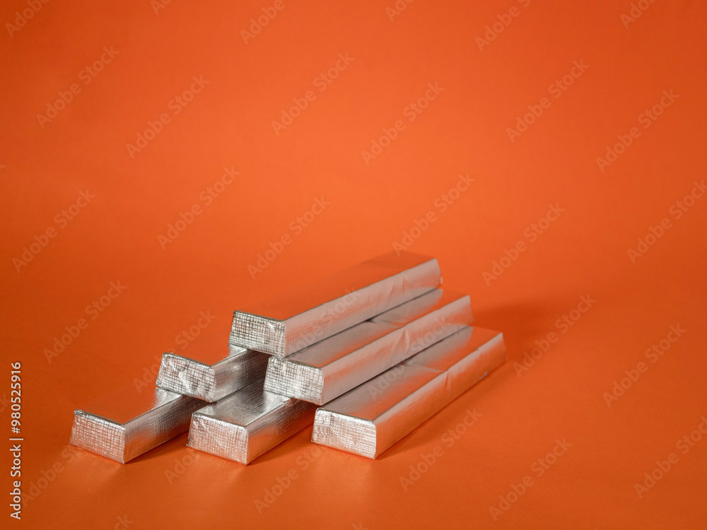 A stack of silver metal bars on a red background. The bars are stacked in a pyramid shape. Invest in precious metals to keep value of money against rising inflation concept. A stack of silver metal bars on a red background. The bars are stacked in a pyramid shape. Invest in precious metals to keep value of money against rising inflation concept.