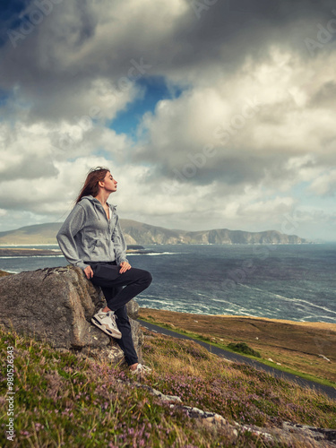 Teenager girl with long hair in grey hoodie is sitting on a rock overlooking stunning nature scenery of Achill island, Ireland.. The scene has a calm and peaceful mood. Travel and tourism concept.