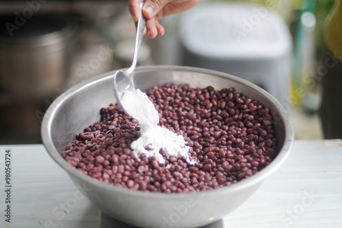 Pouring tempeh or tempe starter (Rhizopus oligosporus,  Rhizopus oryzae) to dried boiled azuki (adzuki) in mixing bowl on white table closed up selective focus blurred background photo