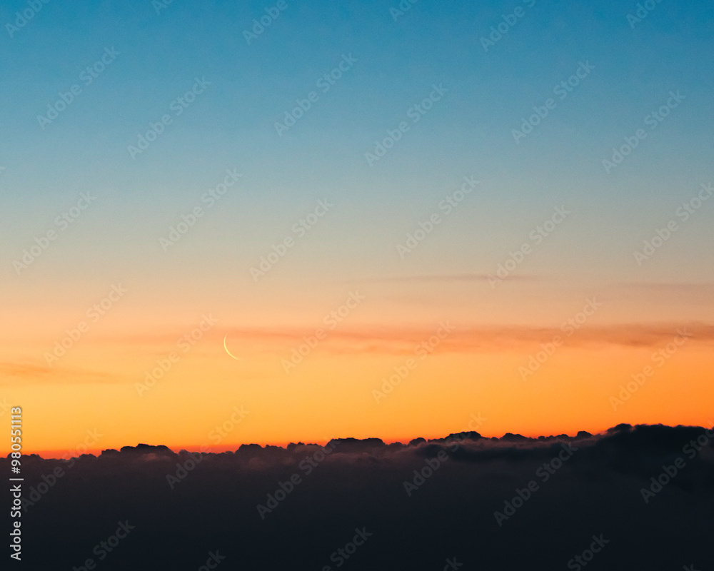 Sunrise and waning moon above black clouds and below clear sky with beautiful colors from blue to orange Sunrise and waning moon above black clouds and below clear sky with beautiful colors from blue to orange