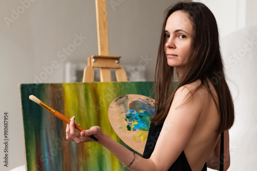 Portrait of emale artist working in studio standing in front of easel with colored canvas with brush and palette in her hands photo