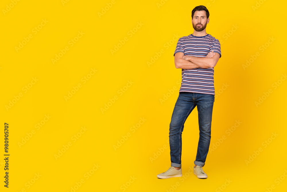 Full body length photo of confident serious bearded man wearing striped t shirt and denim jeans near copyspace isolated on yellow color background Full body length photo of confident serious bearded man wearing striped t shirt and denim jeans near copyspace isolated on yellow color background