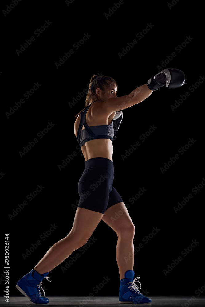 Fototapeta premium Focused young woman with fit body, boxing athlete in motion in gloves, practicing, fighting against black studio background. Concept of sport, hobby, active and healthy lifestyle. Full-length