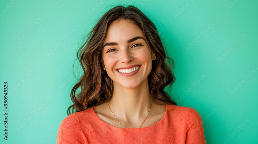 smiling young woman with long, wavy brown hair smiling brightly wearing a coral top against mint green background smiling young woman with long, wavy brown hair smiling brightly wearing a coral top against mint green background