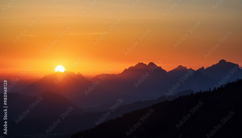 Obraz premium The sun sets behind a distant mountain range, casting warm orange and red hues over the silhouetted peaks and valleys.