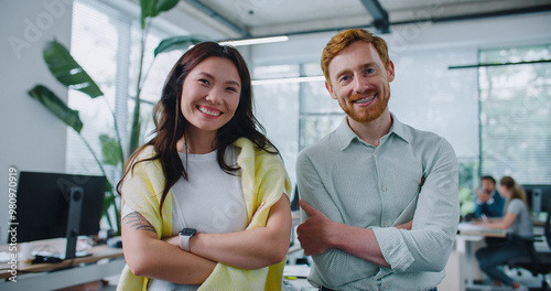 Two people standing side by side and confidently looking directly at camera. Woman with dark hair and yellow sweater over her shoulders positively smiling. Handsome man with red hair crossing his arms