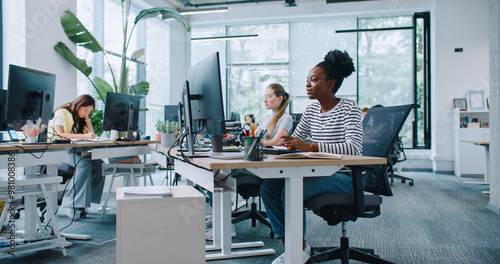 Zooming in on pretty woman with casual clothes working at computer in office. Distracted by camera from work and joyfully smiling. In background people sitting at their workplaces. Job concept.