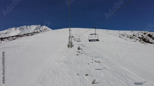 Four person ski chair lift in blue sky going up a mountain with skiers on the piste below
