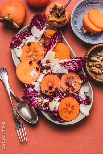 Autumn, winter persimmons salad  with red radicchio Treviso (PGI, Verona, Italy) lettuce, walnuts, pomegranate seeds, aged Parmesan cheese and honey dressing, orange background. Healthy food recipes photo