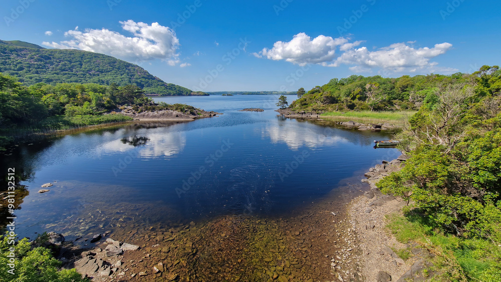 Fototapeta premium A breathtaking view of Muckross Lake under the clear blue sky, where the shimmering waters mirror the lush greenery and surrounding mountains, inviting all to embrace the beauty of nature.
