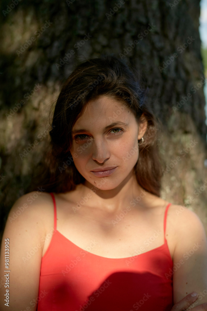 A confident woman in a red dress posing in front of a tree with dappled sunlight illuminating her face. A confident woman in a red dress posing in front of a tree with dappled sunlight illuminating her face.