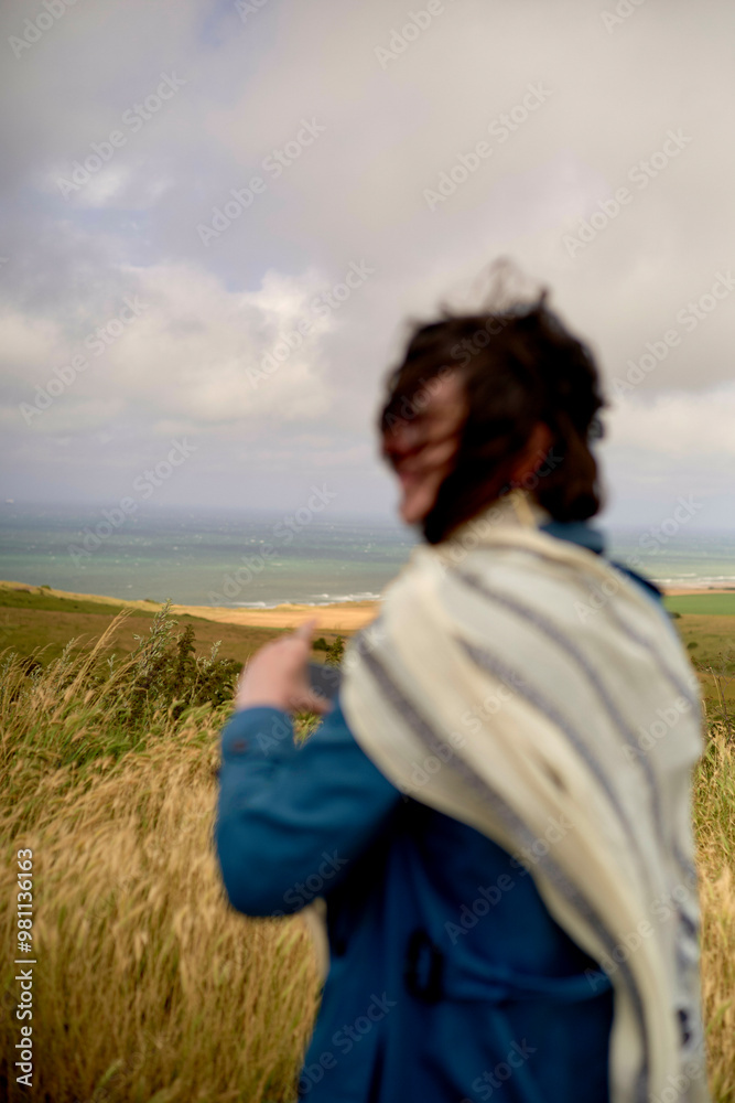 A person stands in a field overlooking the sea, with windblown hair and a scarf draped around their neck. A person stands in a field overlooking the sea, with windblown hair and a scarf draped around their neck.
