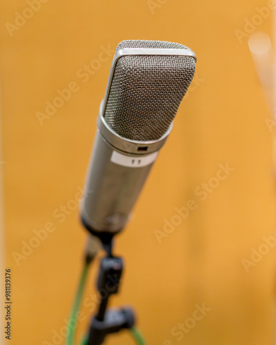 A studio microphone with a silver grille body mounted on a stand, sharp in focus against a blurred orange background. photo