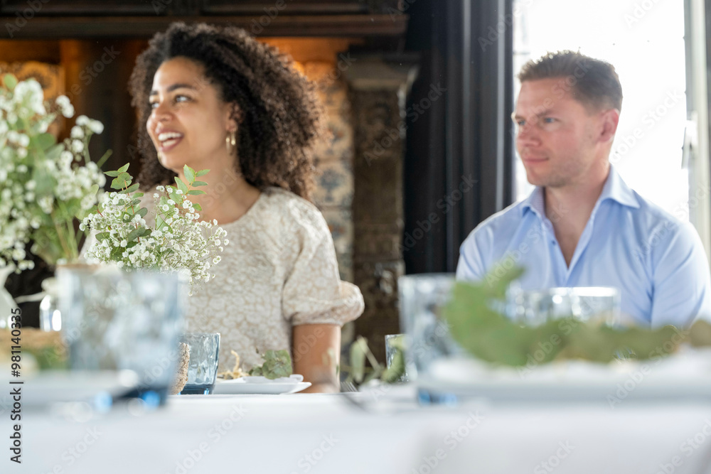 A smiling woman in a white lace dress and a man in a blue shirt are sitting at a table adorned with white flowers during a daytime event. A smiling woman in a white lace dress and a man in a blue shirt are sitting at a table adorned with white flowers during a daytime event.