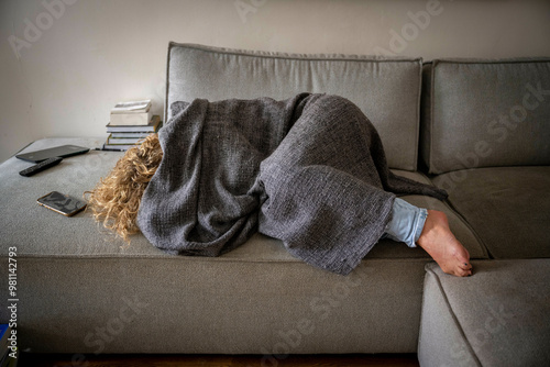 Adult person napping on a grey couch covered with a dark blanket, their foot peeking out, next to a smartphone and a stack of books. photo