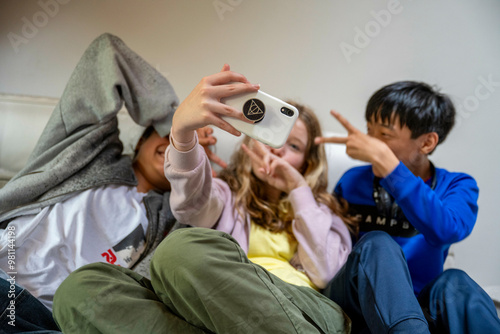 Three kids take a selfie together, one covering their face with a hoodie, another making a peace sign, and the third smiling at the camera.