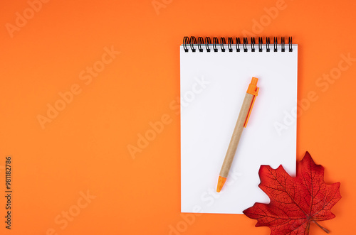Flatlay composition with blank notepad, pen and autumn leaf on orange background. Top view. Copy space.