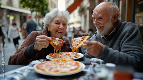 A joyful elderly Caucasian couple sharing pizza slices outdoors, enjoying a relaxed meal together on a city street, smiling.