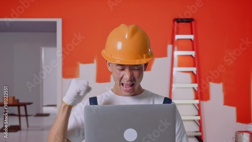 Close Up Of Asian Man Painter Wearing Safety Helmet Looking At The Laptop Then Screaming Goal Celebrating While Standing With Painting Wall In Room photo