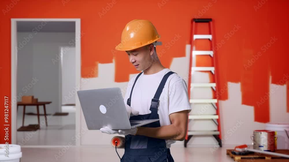 Side View Asian Man Painter Wearing Safety Helmet Using A Laptop While Standing With Painting Wall In Room Side View Asian Man Painter Wearing Safety Helmet Using A Laptop While Standing With Painting Wall In Room