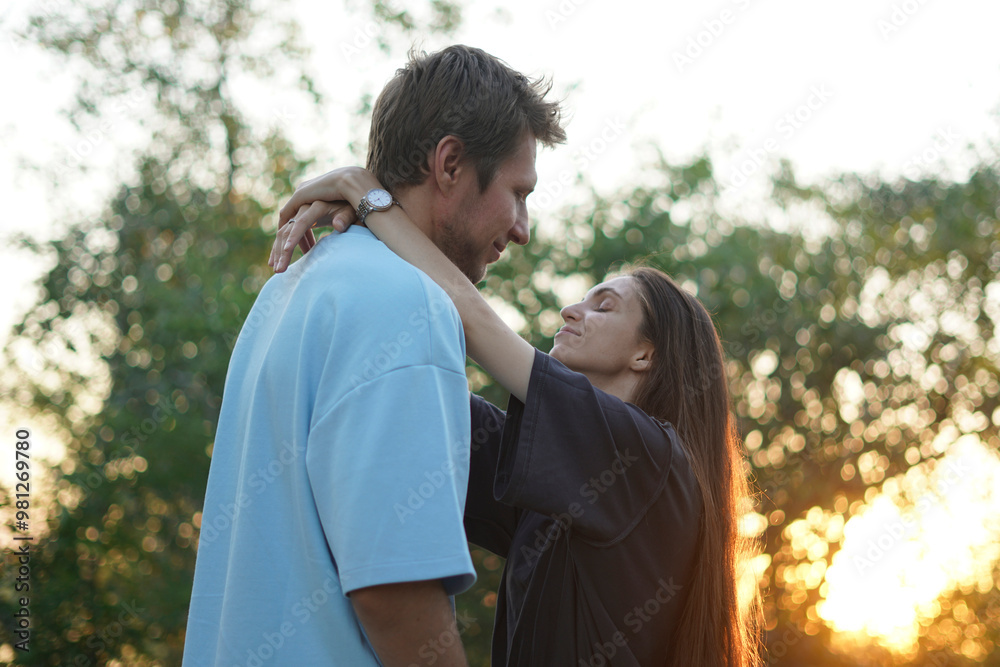 Fototapeta premium woman in a black shirt embraces a man in a blue shirt as they stand in a forest. The sun shines behind them, creating a warm, golden glow