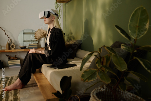 Side view wide shot of young girl with long white hair sitting on couch in wabi sabi living room watching movie via VR headset photo