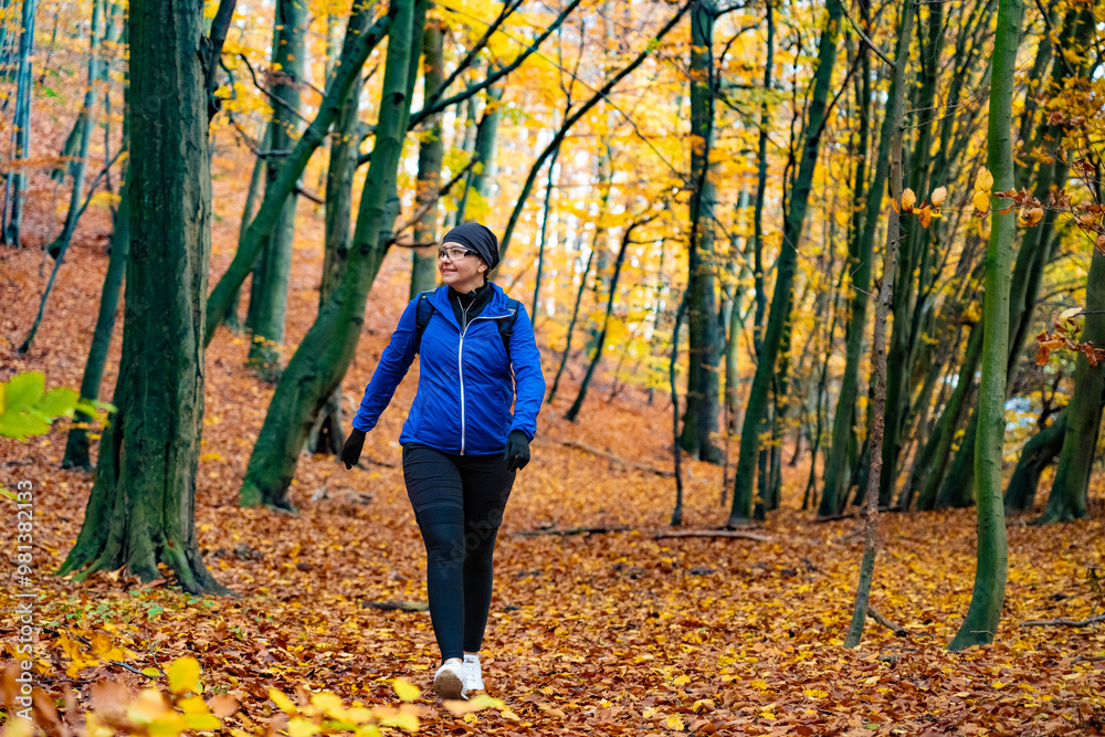 Middle-aged woman dressed sporty in blue jacket and black pants, cap and glasses training in forest full of fallen leaves in autumn weather. Front view Middle-aged woman dressed sporty in blue jacket and black pants, cap and glasses training in forest full of fallen leaves in autumn weather. Front view