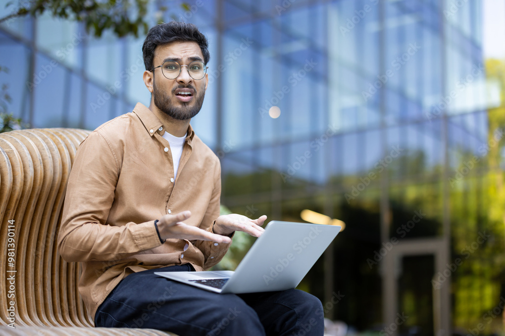 Man sitting on bench using laptop outdoors. Expresses confusion or frustration with work or study. Background features modern glass building. Casual attire suggests relaxed work or study environment. Man sitting on bench using laptop outdoors. Expresses confusion or frustration with work or study. Background features modern glass building. Casual attire suggests relaxed work or study environment.