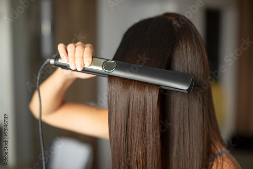 Konstfotografi Young brunette woman straightening her glowing healthy hair using modern iron ha