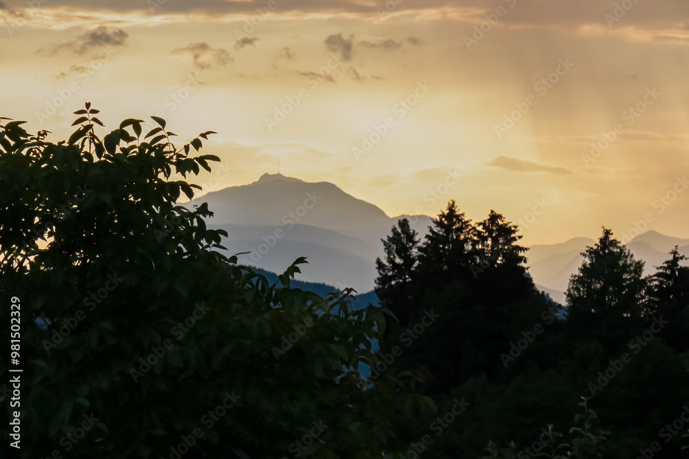 Obraz premium Fiery sunset over mountain summit Dobratsch in Rosental, Carinthia, Austria, Europe. Silhouette of forest and rolling hills in Austrian Alps. Vibrant orange and yellow hues. Tranquil serene atmosphere