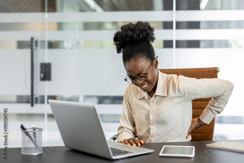 African american woman feeling back discomfort . Seated desk with laptop, illustrating workplace health challenges. Expresses stress or strain associated with office work and prolonged computer use.