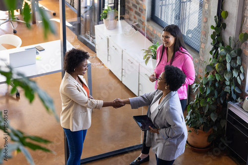 Businesswomen shaking hands in modern office, colleague holding tablet and smiling