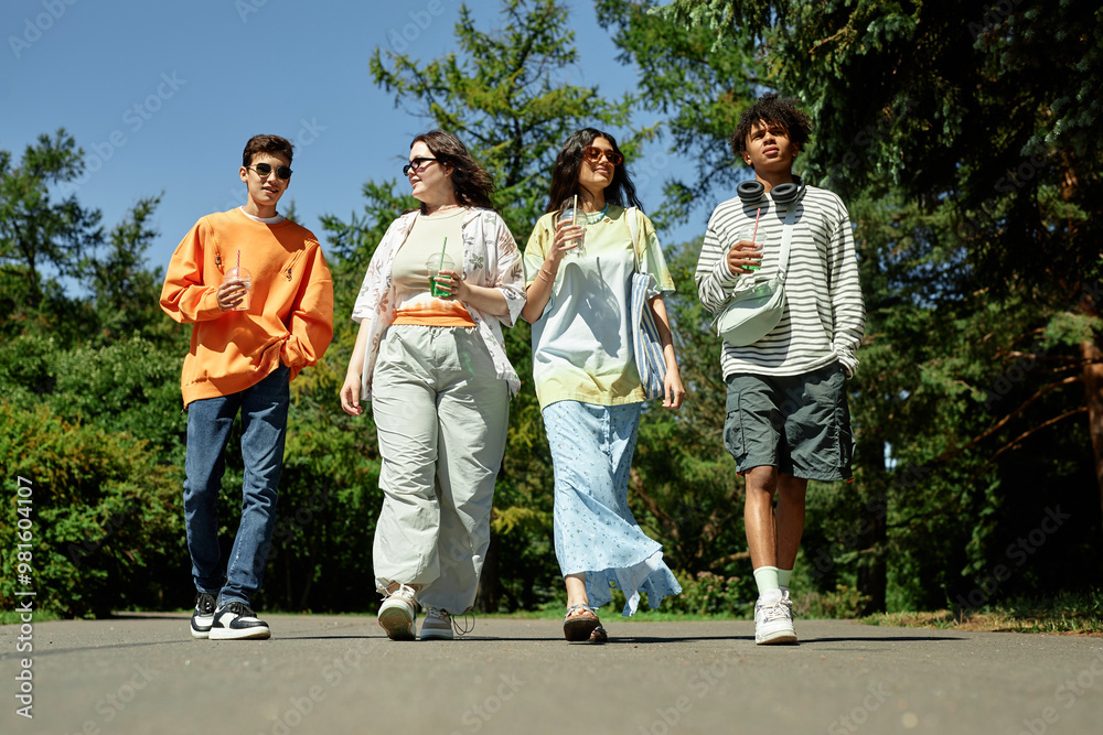 Full length shot of four teenagers chatting while walking along park path enjoying sunny weather and refreshing drinks Full length shot of four teenagers chatting while walking along park path enjoying sunny weather and refreshing drinks