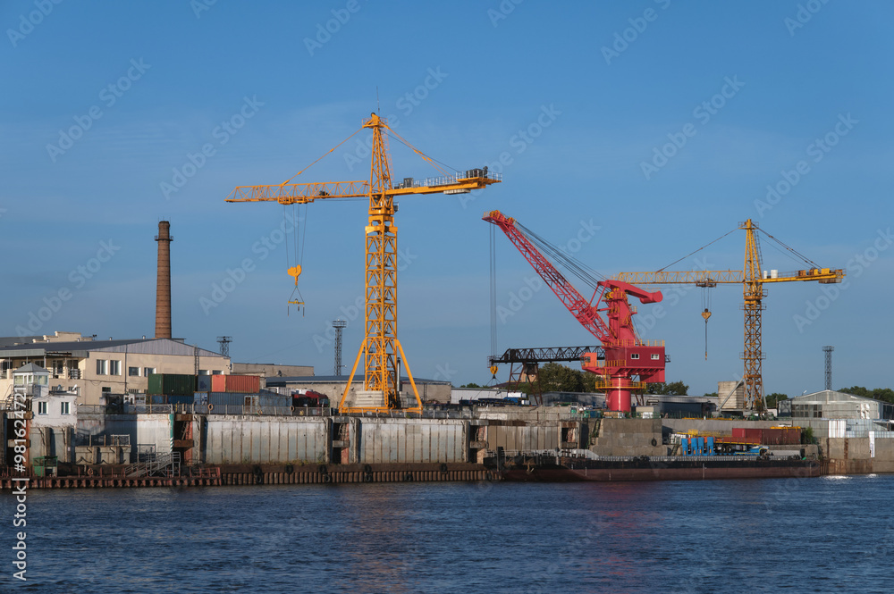 Port cranes on the pier of the commercial port on a summer evening. Bright colors in the rays of the setting sun. The border river Amur between Russia and China. Port cranes on the pier of the commercial port on a summer evening. Bright colors in the rays of the setting sun. The border river Amur between Russia and China.