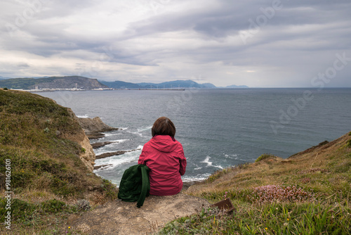 Caucasian woman with red jacket sitting by the cliffs and sea waves, Basque Country, Spain.