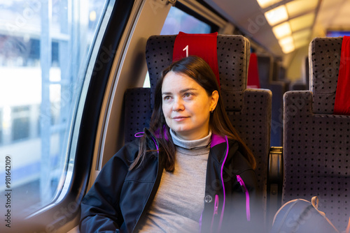 Dreamy dark-haired woman enjoying journey in modern comfortable train, looking at picturesque scenery outside window.. photo