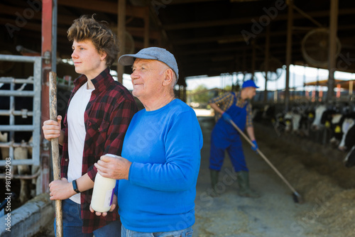 Positive aged man dairy farm owner standing with teen grandson near stalls with cows on sunny summer day, holding bottle of fresh milk .. photo