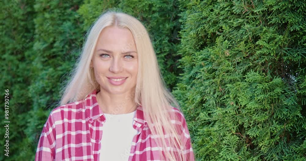 Portrait of attractive female gardener with blond hair wearing casual clothes standing near green thujas. Young positive woman posing near hedge at garden. Portrait of attractive female gardener with blond hair wearing casual clothes standing near green thujas. Young positive woman posing near hedge at garden.
