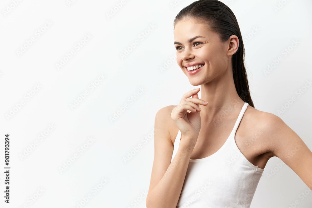 Smiling young woman with long hair wearing a white tank top, posed against a neutral light background, conveying joy and confidence with an expressive gesture Smiling young woman with long hair wearing a white tank top, posed against a neutral light background, conveying joy and confidence with an expressive gesture