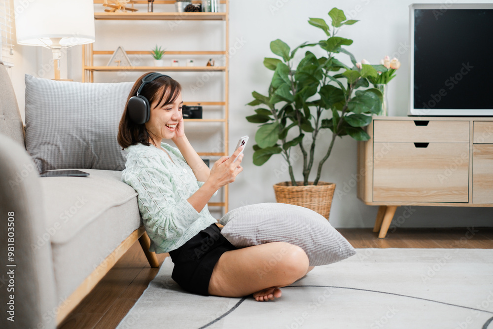 Happy young woman enjoying music on headphones while using her smartphone in a cozy, modern living room. Happy young woman enjoying music on headphones while using her smartphone in a cozy, modern living room.