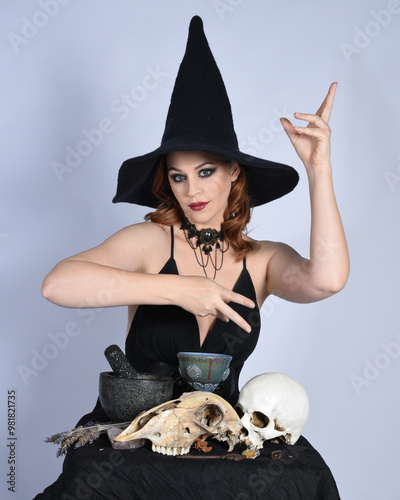 Close up portrait of beautiful  female model wearing black dress, pointy witch hat halloween costume.  Isolated figure on studio background. Casting magic spell at spooky occult  ritual potion table  photo