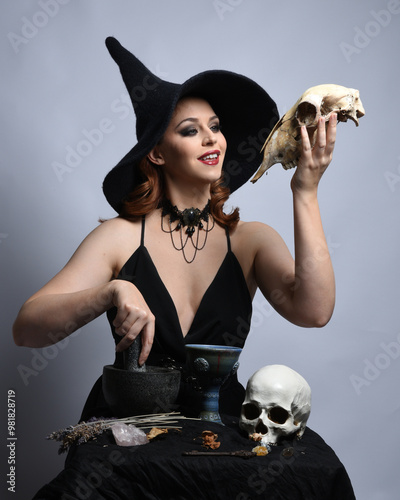 Close up portrait of beautiful  female model wearing black dress, pointy witch hat halloween costume.  Isolated figure on studio background. Casting magic spell at spooky occult  ritual potion table  photo