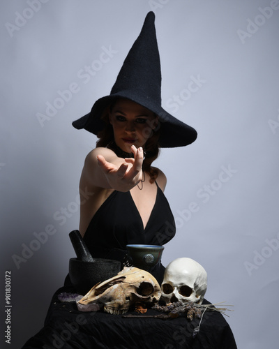 Close up portrait of beautiful  female model wearing black dress, pointy witch hat halloween costume.  Isolated figure on studio background. Casting magic spell at spooky occult  ritual potion table  photo