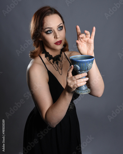 Close up portrait of beautiful  female model wearing black dress, pointy witch hat halloween costume.  Isolated figure on studio background. Holding wine goblet at spooky occult ritual potion table  photo