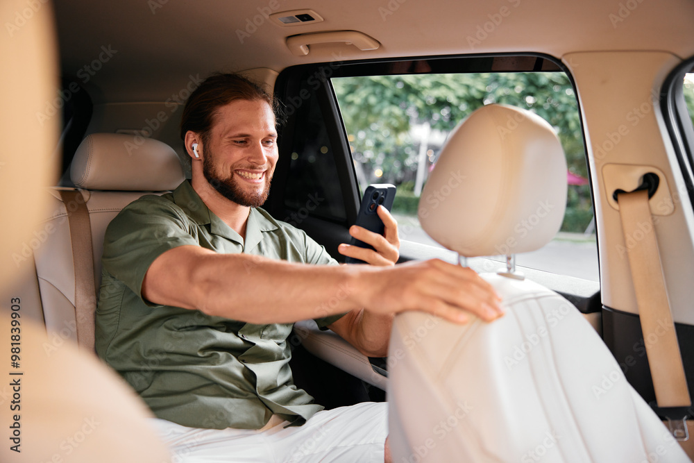 Smiling man sitting in the backseat of a car, using a smartphone while wearing wireless earbuds
