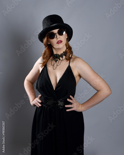 Close up portrait of beautiful female model wearing black dress and steampunk spectacles and top hat halloween costume. Isolated figure on studio background.