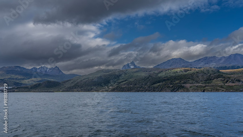 The picturesque coastline of Argentina. Snow-capped Andes mountains against the sky, clouds. At the foot of the hills are the city buildings of Ushuaia. Ripples on the ocean surface. Tierra del Fuego