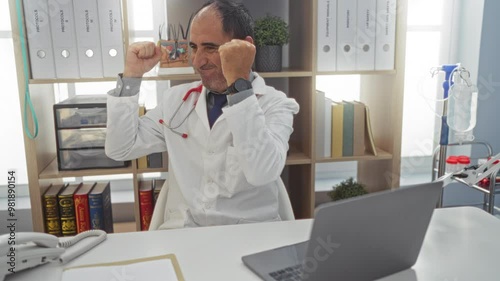 Wallpaper Mural A middle-aged hispanic male doctor works on a laptop in a clinic office, appearing both concentrated and jubilant, surrounded by medical equipment and documentation. Torontodigital.ca